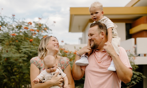 A smiling family stands outdoors near greenery and a building, with a father holding a young child on his shoulders while a mother holds a baby in her arms.