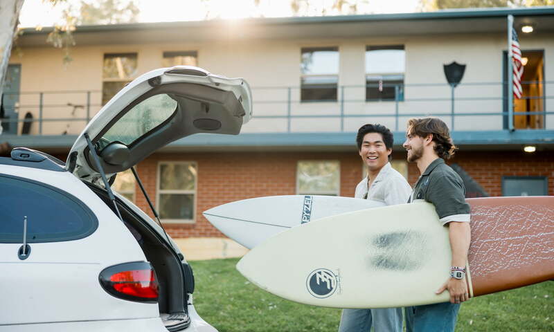 two students load their surfboard into a parked car in front of Stewart Hall 