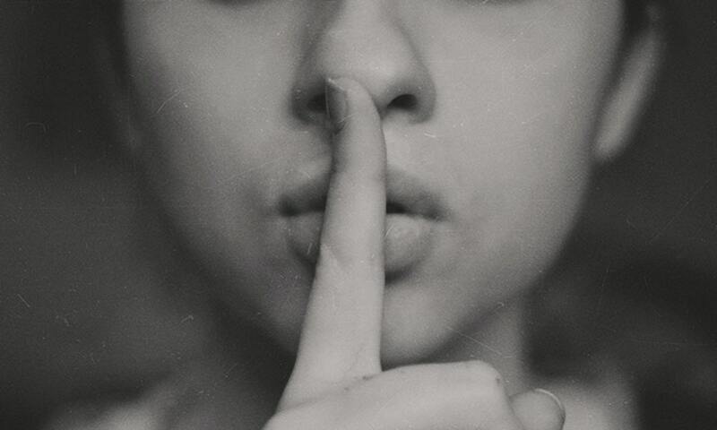 Image shows a women putting her finger in front of her lips as if to tell someone to be quiet
