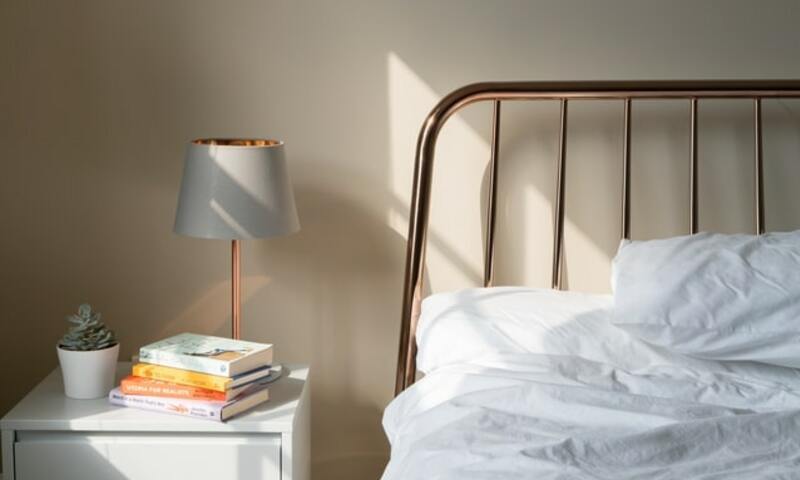 Image shows a bed and bedside table with books and a lamp in daylight. 