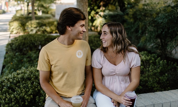 A young couple sits together outdoors, smiling at each other while holding drinks in a peaceful garden setting.