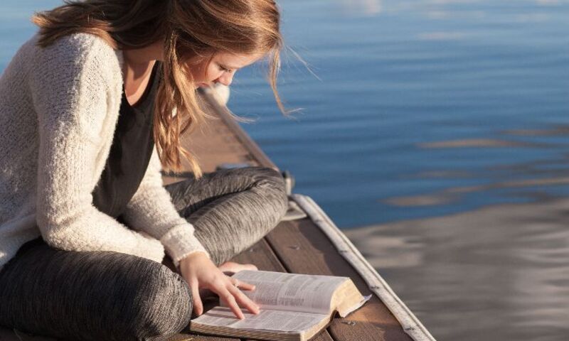 Woman sitting at end of dock overlooking lake while studying the Bible