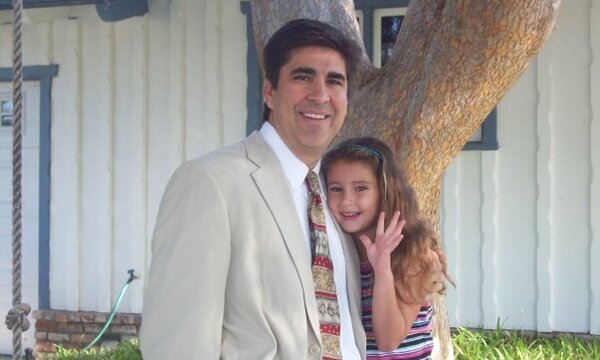Dr. Chris Grace stands beside his young daughter, Caroline, smiling outdoors near a large tree.