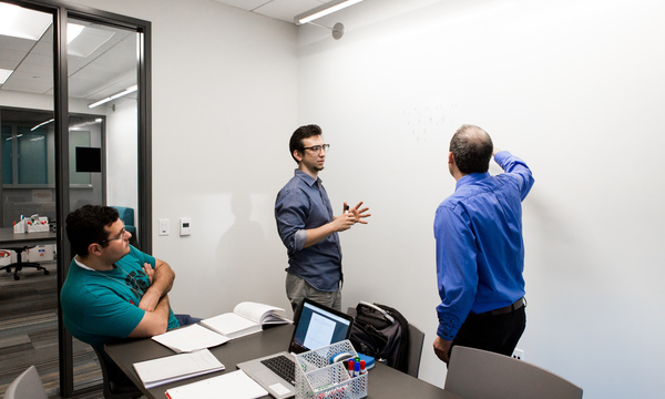 Three young men talking in a room with a table and whiteboard