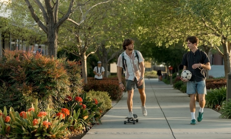 Two students, one on a skateboard, travel down the path in front of the library. 