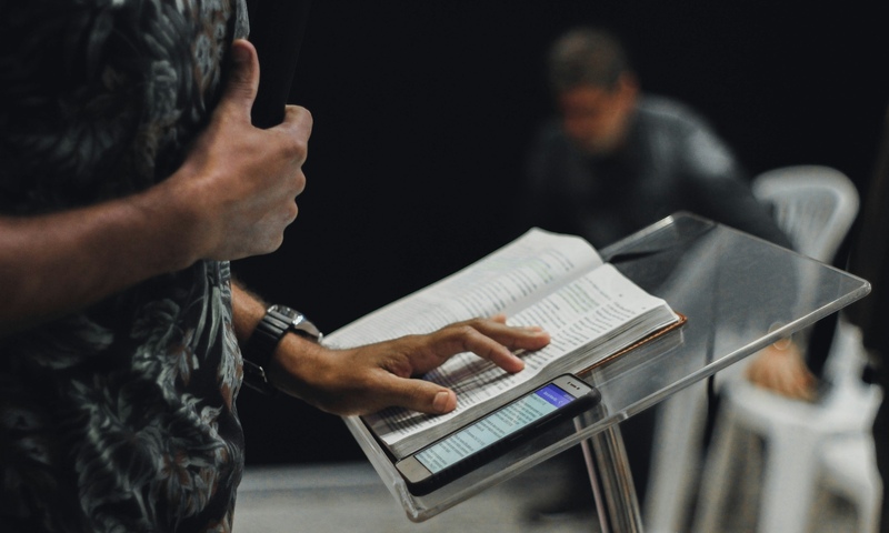 image shows a Bible open on a stand with a hand on it as if preaching 
