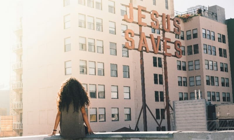 Girl sits at top of Ace Hotel in Los Angeles overlooking "Jesus Saves" sign