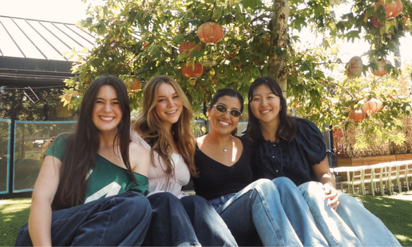 Four young women sit together outdoors, smiling at the camera beneath a leafy tree decorated with hanging lanterns. Sunlight filters through the branches, creating a warm and welcoming atmosphere.