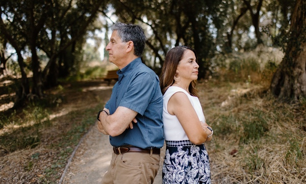 A husband and wife stand back to back with folded arms on a wooded path, looking in opposite directions, illustrating tension or disagreement.