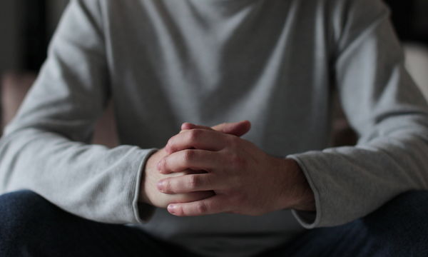 Person sitting with hands clasped and head out of frame, wearing a light gray long-sleeve shirt in a dimly lit room.