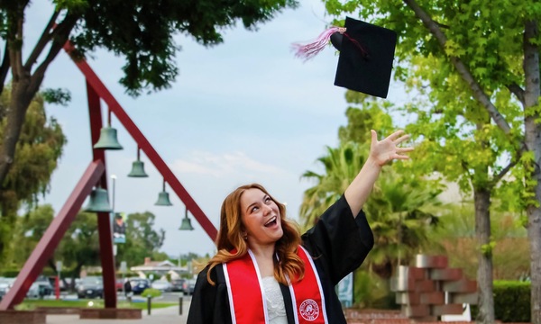 Graduate in a black cap and gown with a red “Class of 2025” Biola University stole tosses her cap in the air, smiling excitedly on a tree-lined campus walkway with bells and greenery in the background.