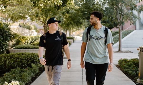 Two college students walking together and talking on a tree-lined campus walkway.
