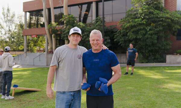 Father and son standing together on a college campus lawn, smiling with their arms around each other during an outdoor gathering.