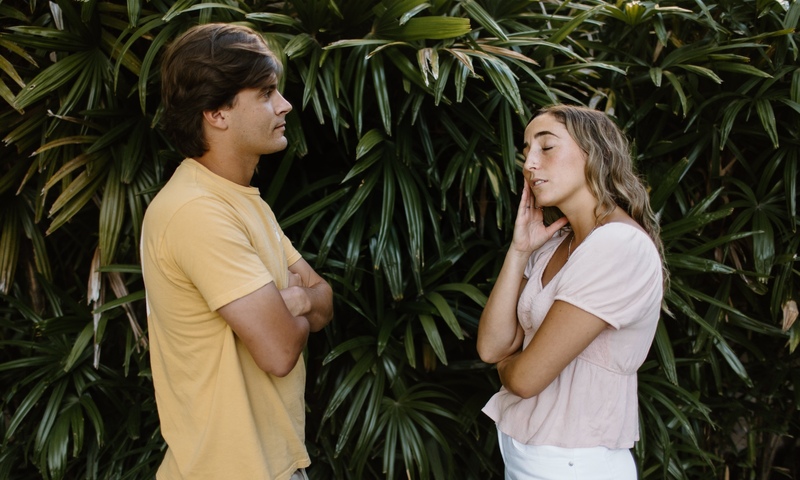 A young couple stands facing each other outdoors. The man, wearing a yellow T-shirt, has his arms crossed and looks serious. The woman, dressed in a light pink blouse and white pants, touches her face with her eyes closed, appearing frustrated or upset.