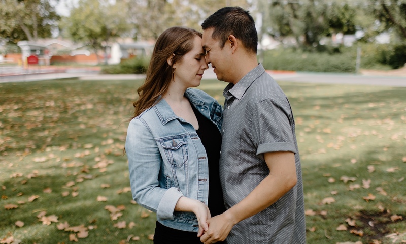 A couple stands closely together in a park, gently holding hands with their foreheads touching.