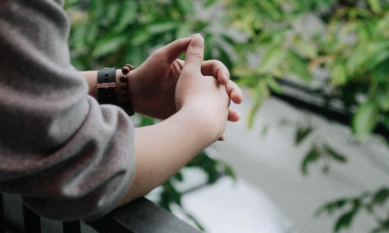 Hands folded in prayer over railing with greenery in background
