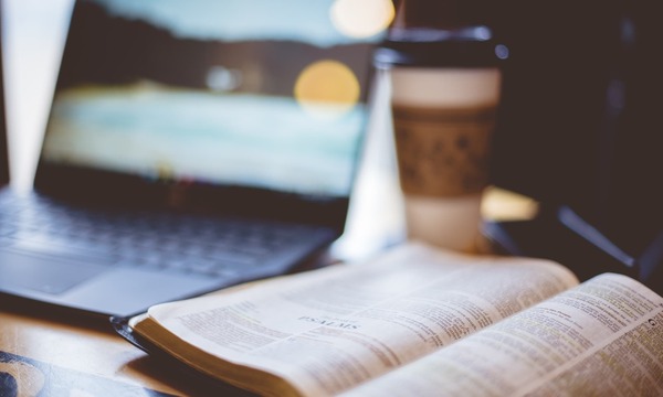 image shows a Bible with a computer open in the background