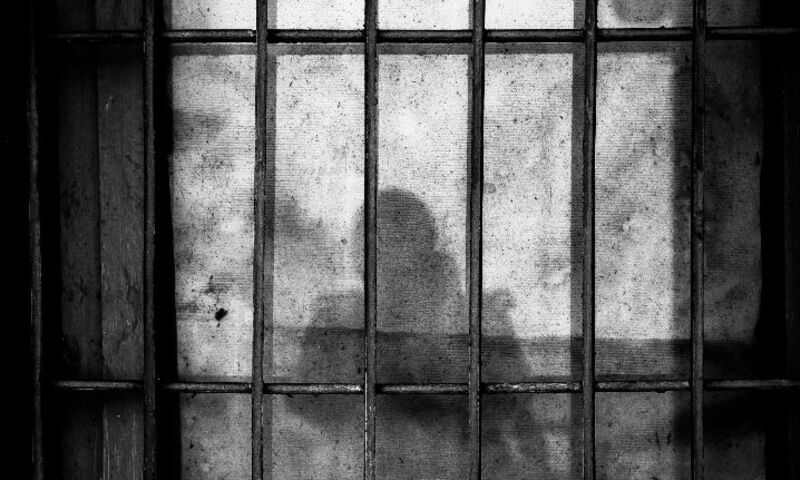 Image shows blurred shadow of person behind prison bars in black and white