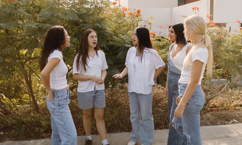 Five college-aged women standing together talking outside on a sunny campus walkway.