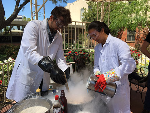 two students in white lab coats holding instruments next to a bowl emitting gas