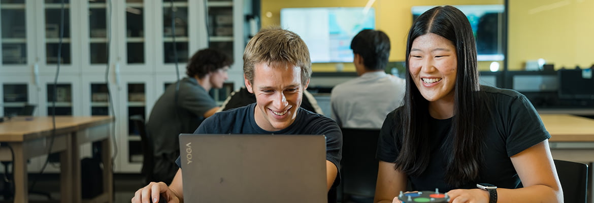 Two students sit together smiling as they work in front of a laptop