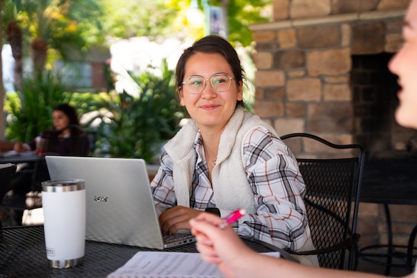 A pair of students sitting outside, talking with each other while doing work. 