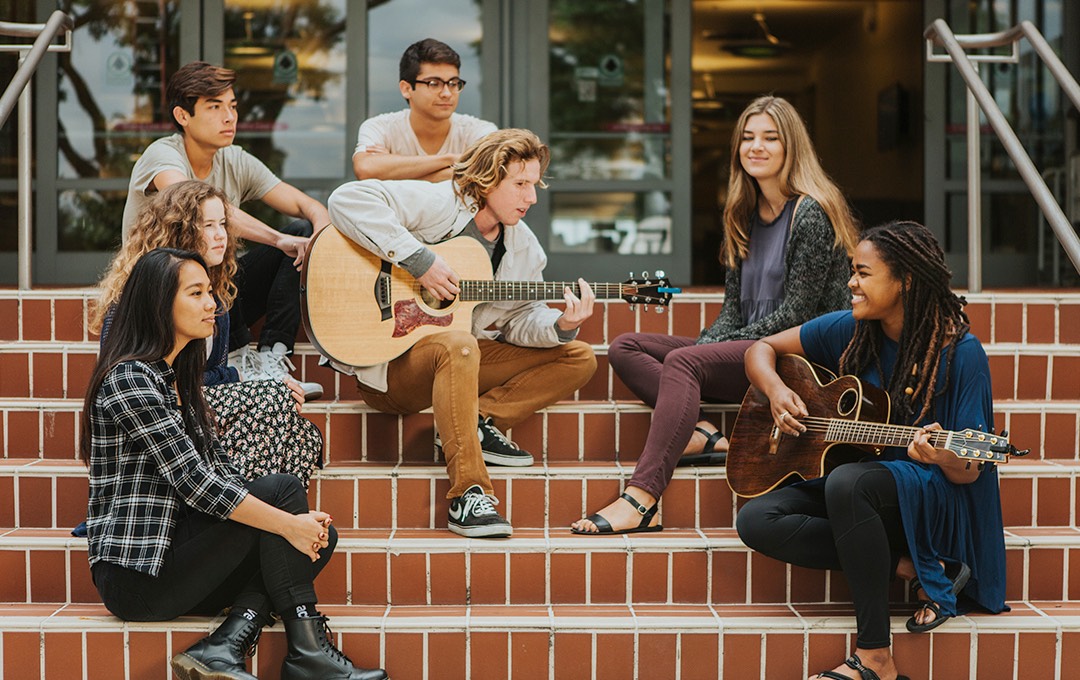 A diverse group of students plays guitar and sings on the steps of the library.