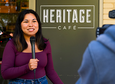 Student with a microphone recording a news segment in front of Heritage Café
