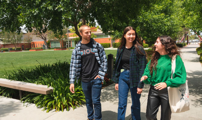 Three students walking outside near trees and a grassy lawn