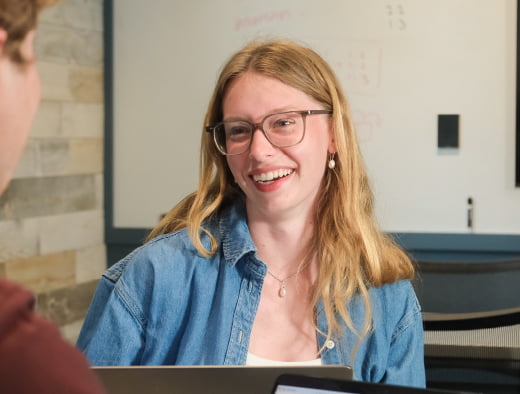 Female student sitting in front of a laptop smiling at someone.
