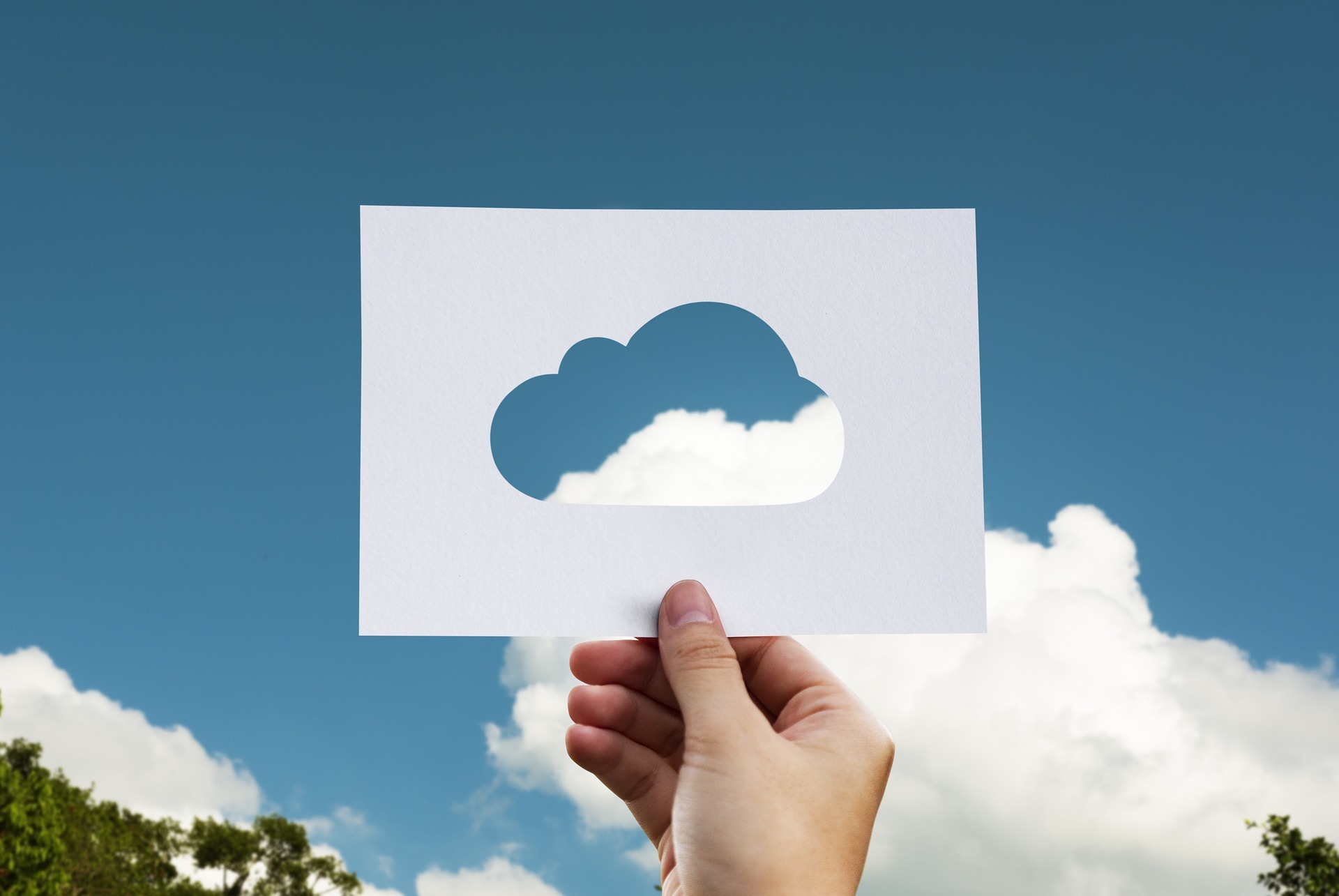 A hand holds a paper up to the sky with a cloud shape cut out.