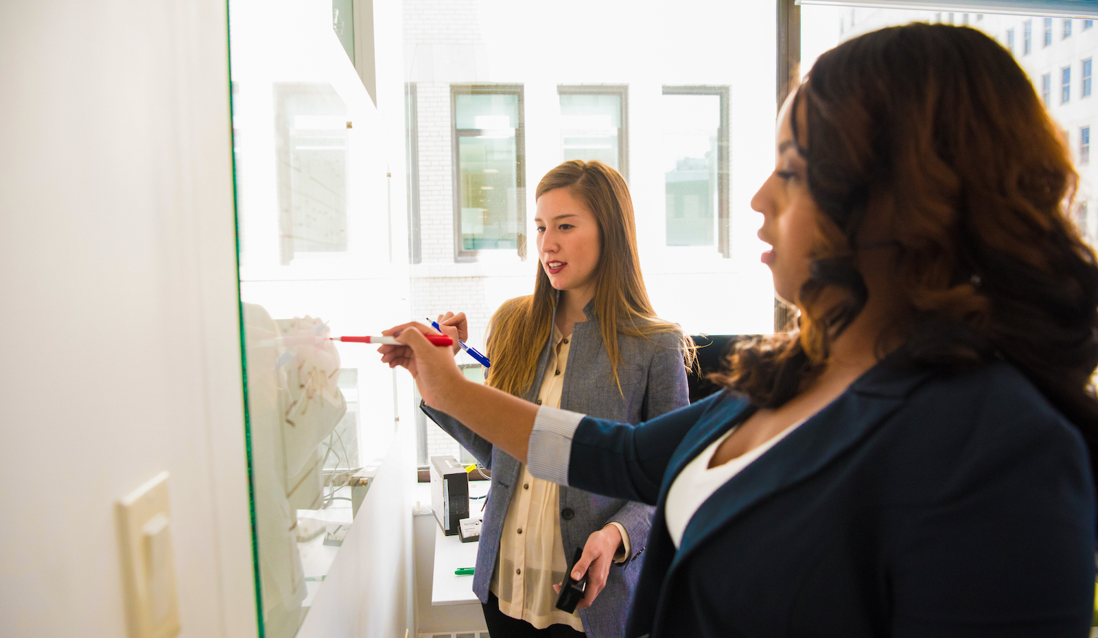 Two women write on a white board.