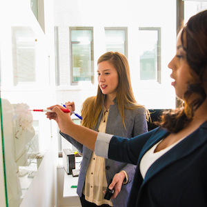 Two women write on a whiteboard.