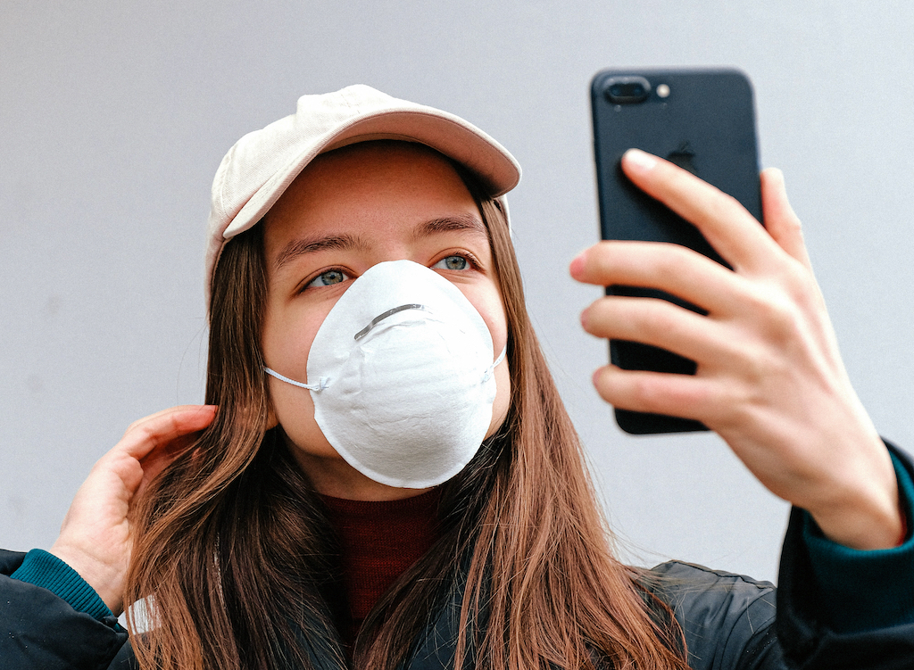 A woman with a dust mask looks at her upheld smartphone as if taking a selfie.