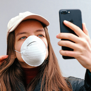 A woman with a dust mask looks at her upheld smartphone as if taking a selfie.