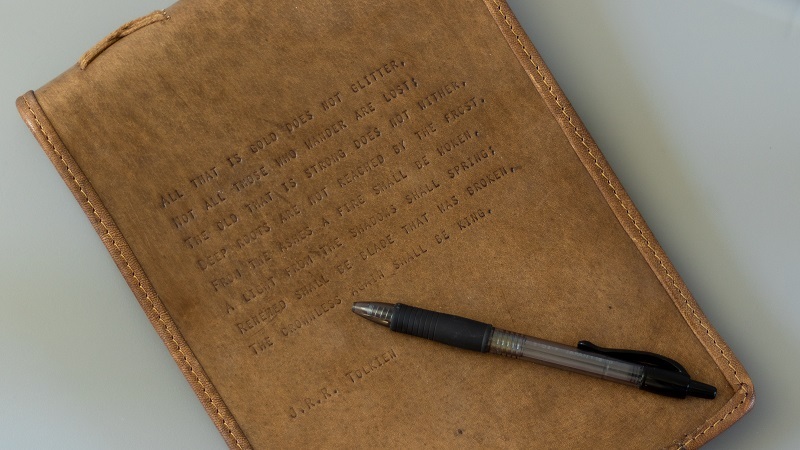 A walnut-stained leather-bound journal with a PILOT G-2 07 pen on top. The quote embossed on the journal's page is J.R.R. Tolkien's "Not all who wonder are lost" quote. See Google for the longer version.