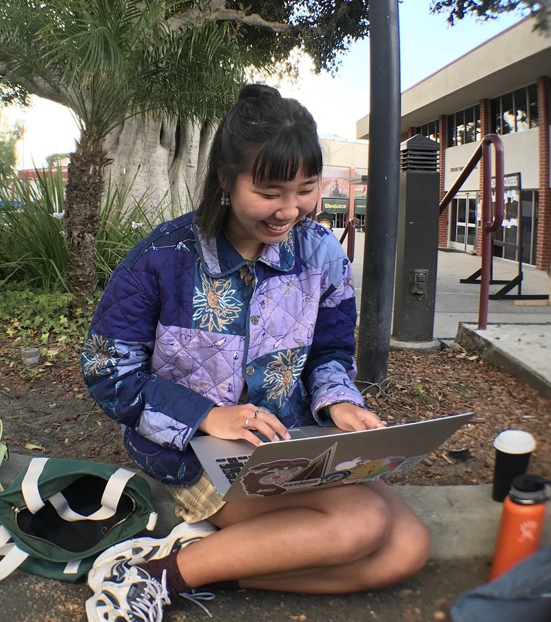 Kristen sitting on the curb of Biola's campus next to the Fireplace Pavilion, waiting in line for Missions Conference. She is wearing a celestial-themed puffy jacket, many rings and at present typing on her laptop