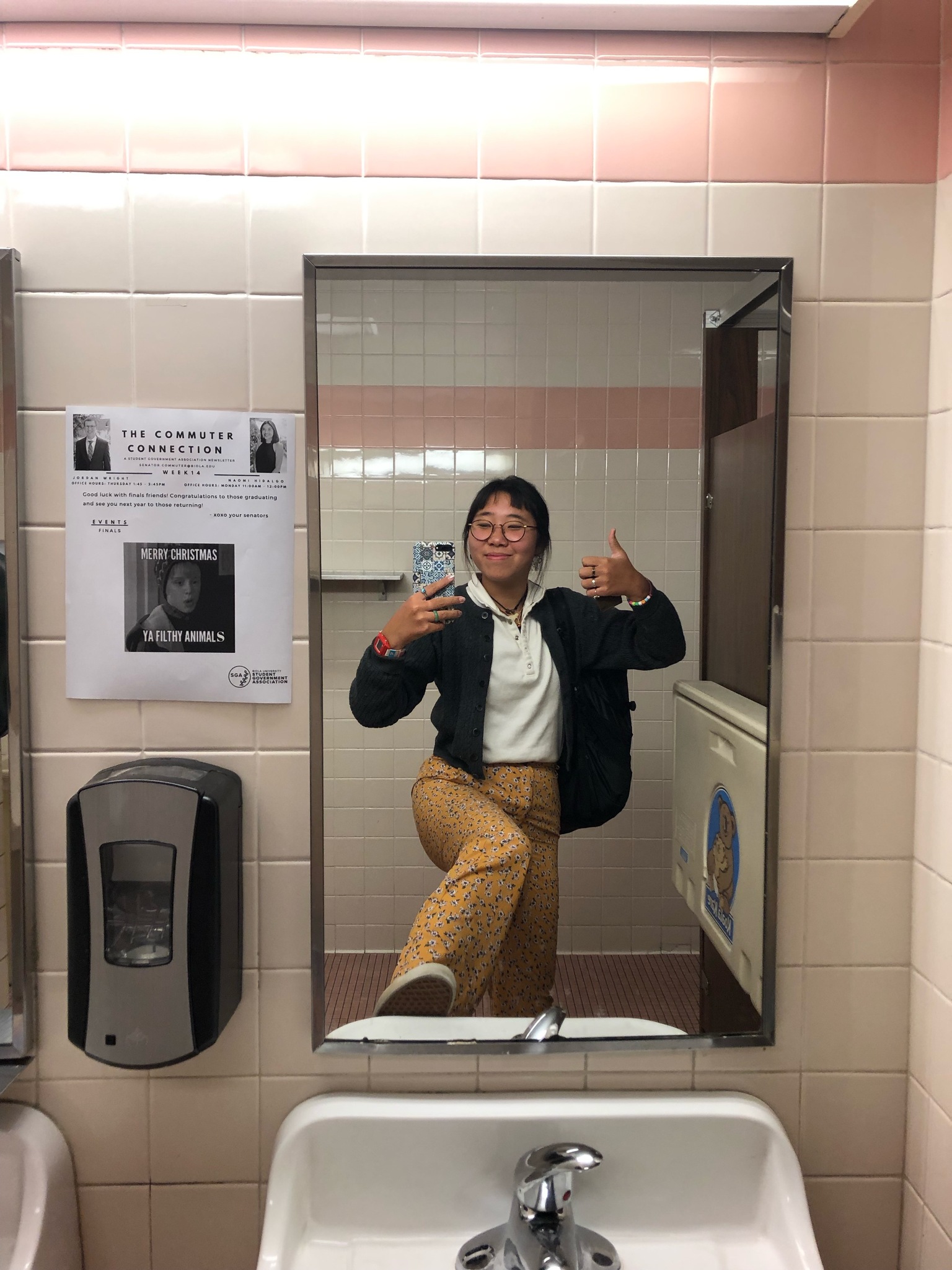 Kristen takes a selfie in one of the campus bathrooms, there's a poster to the side of the mirror detailing a Merry Christmas message for Commuters to campus