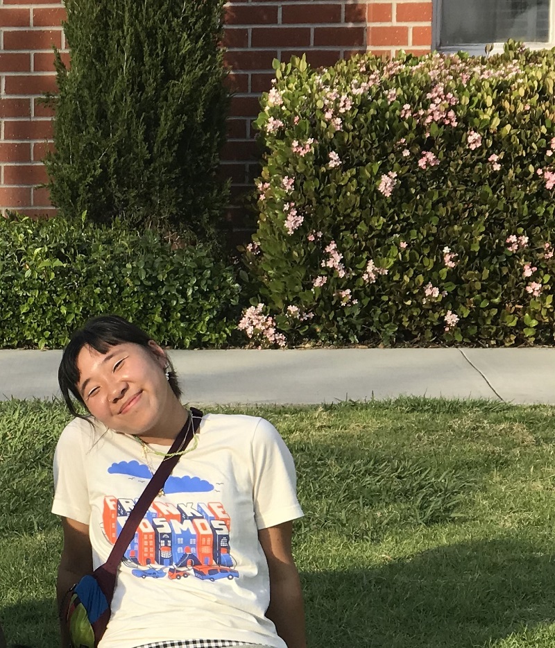 Kristen's sitting on a patch of green by one of 51's brick buildings, waiting for her brother-in-law's senior recital. She's wearing a cream-colored shirt and a fanny pack cross-wise.