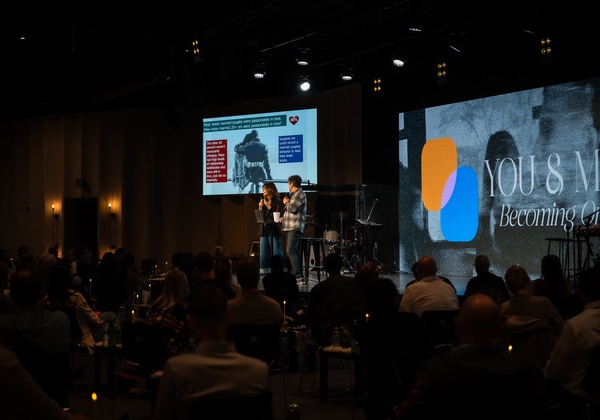 Chris and Alisa Grace speak onstage to an audience during a marriage conference, with presentation slides displayed behind them.