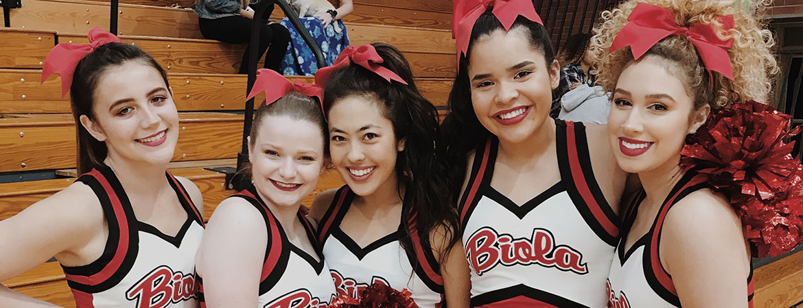 A group of Biola cheerleaders smile together in the gym