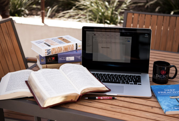 A laptop, books, bible, and mug sit on an outdoor table