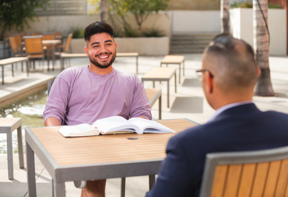 student with books seated outside talking with professor